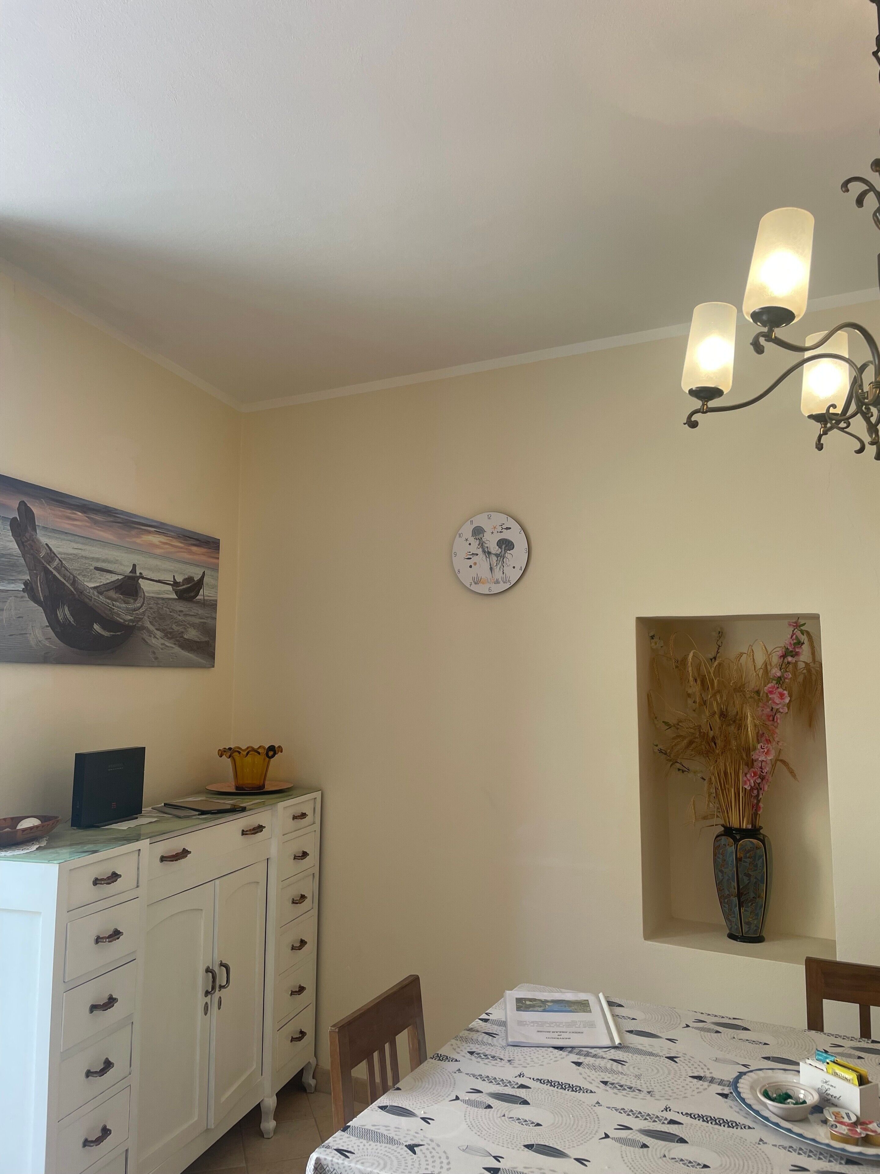 Uma área de jantar com uma mesa posta para dois, um relógio na parede e um quadro sobre os armários.