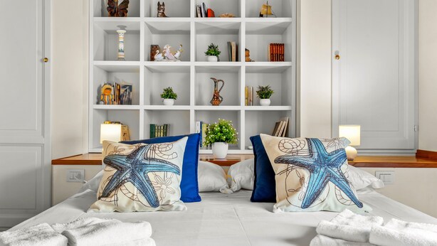 A neatly made bed with starfish pillows, a white nightstand with a lamp, and a white shelving unit with various decorative items.