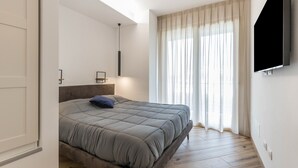 Ein modernes Schlafzimmer mit einem Bett, einem an der Wand montierten Fernseher und einem großen Fenster mit Vorhängen.