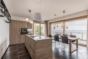 A modern kitchen with wooden cabinets, a central island, and a dining area with a view of the sea.