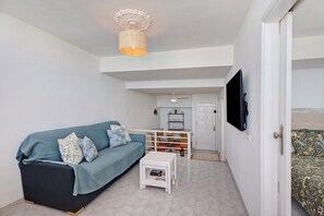 A living room with a sofa, a small table, a TV mounted on the wall, and a door leading to another room.