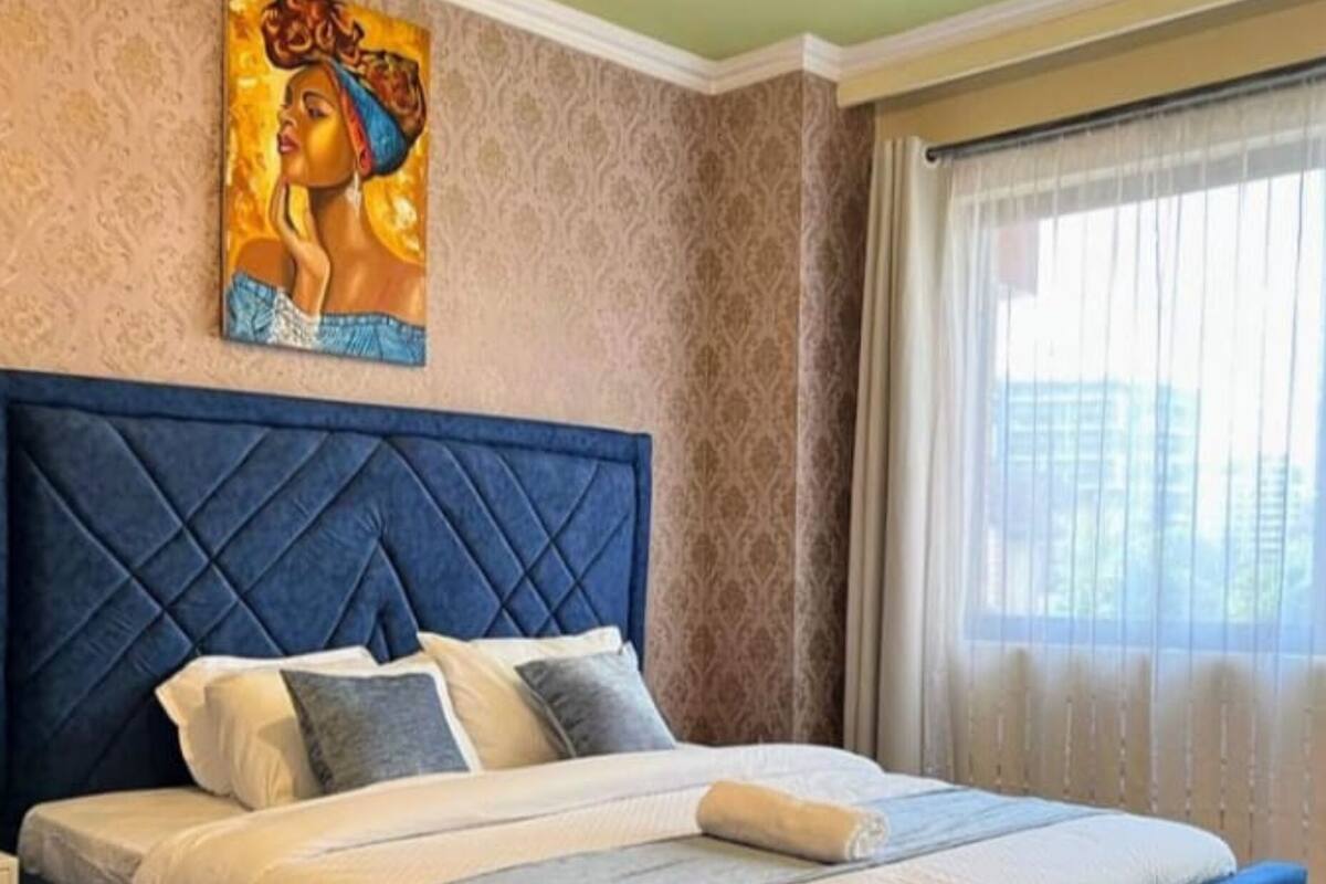 Une chambre à coucher avec un grand lit, une tête de lit, un tableau au mur et une fenêtre avec des rideaux.