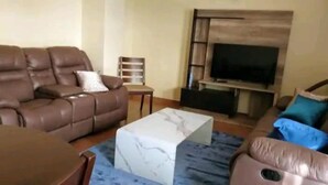 Uma sala de estar com sofá de couro marrom, uma mesa de centro branca, um rack para TV e um tapete azul estampado.