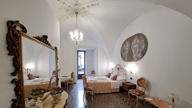 A room with a large ornate mirror, a chandelier, a bed with a headboard, a table with a vase of flowers, and a patterned floor. - Terra d'Amuri Hotel (Catania)