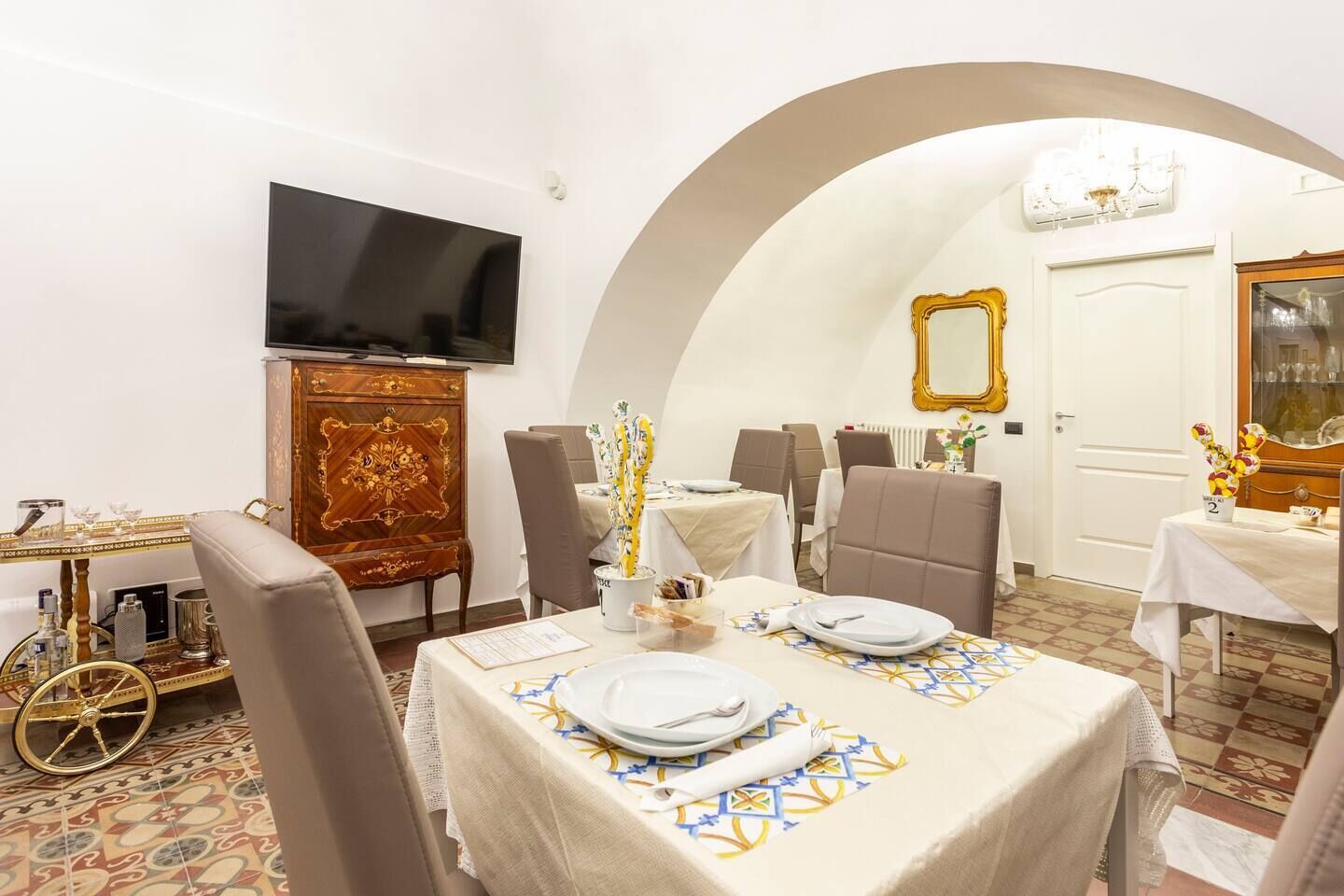 Uma área de refeições com uma mesa posta para dois, uma televisão na parede e um aparador decorativo.