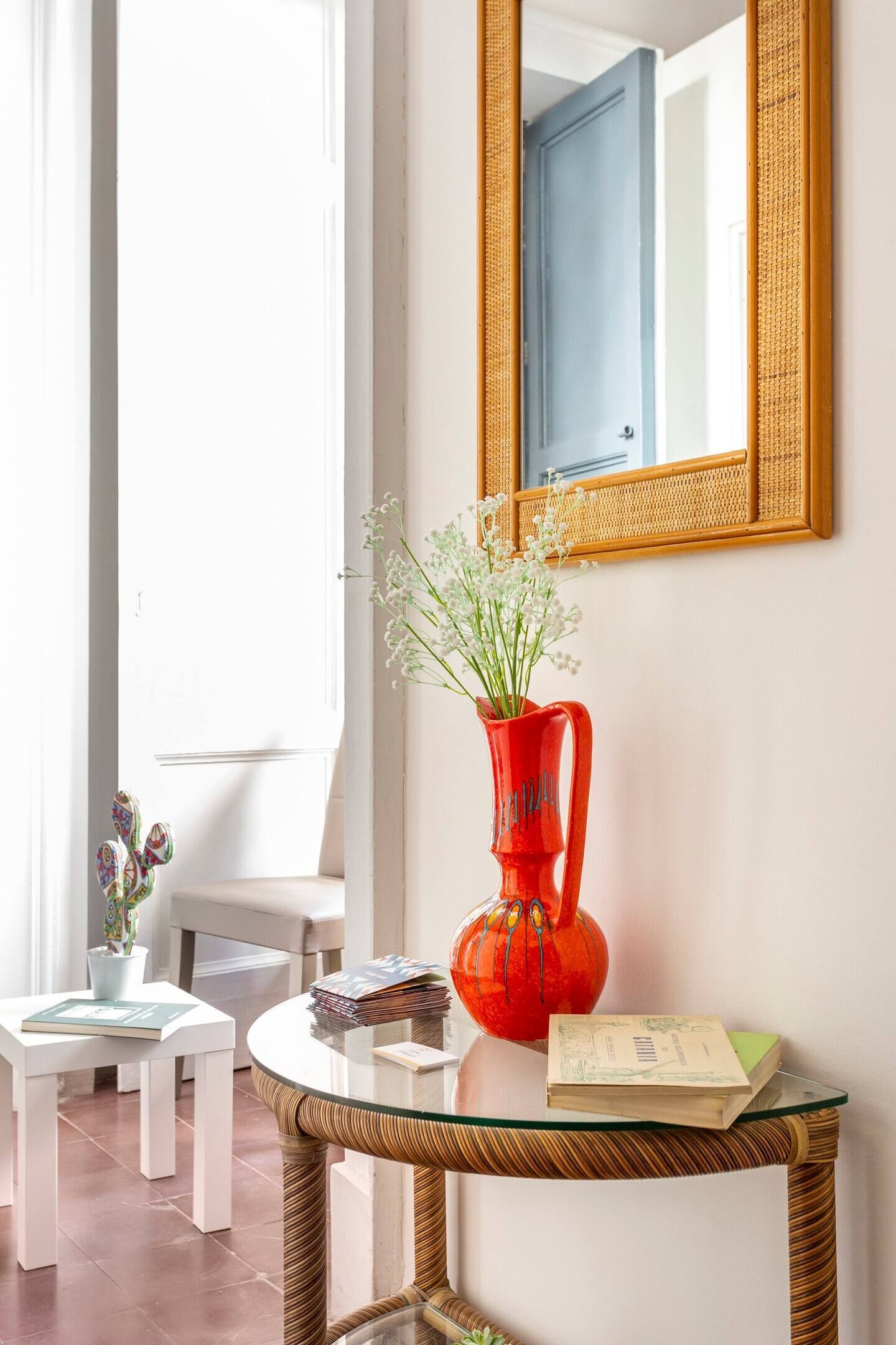 A room with a glass table, a red vase with flowers, a mirror, and a white chair.