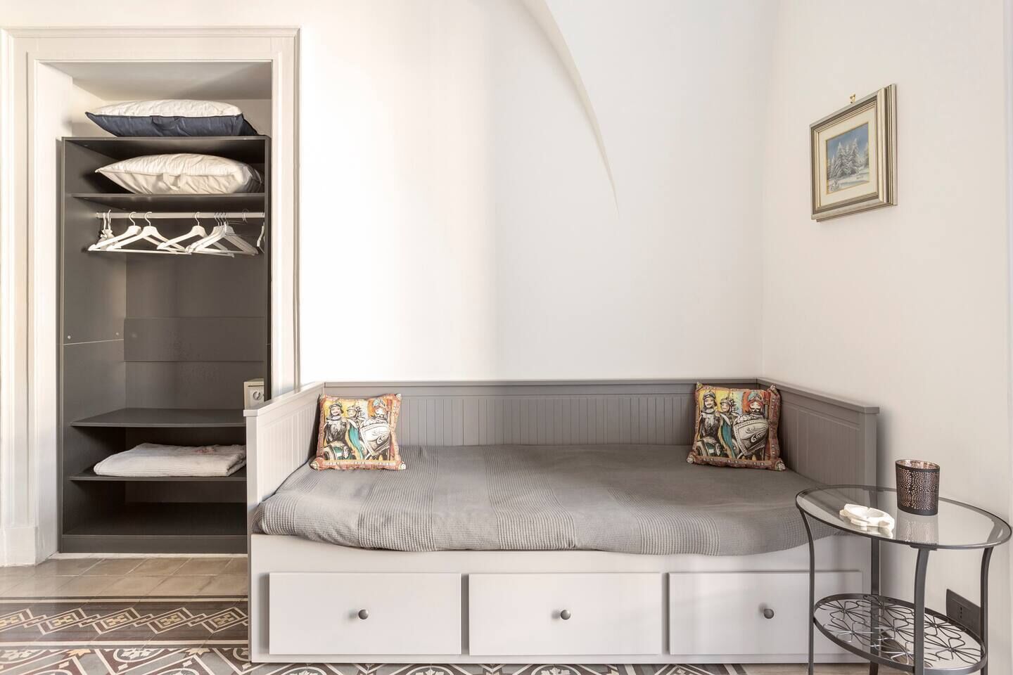 Quarto moderno com sofá cinzento, duas almofadas decorativas, uma pequena mesa de apoio e um guarda-roupa com espaço para pendurar roupas.