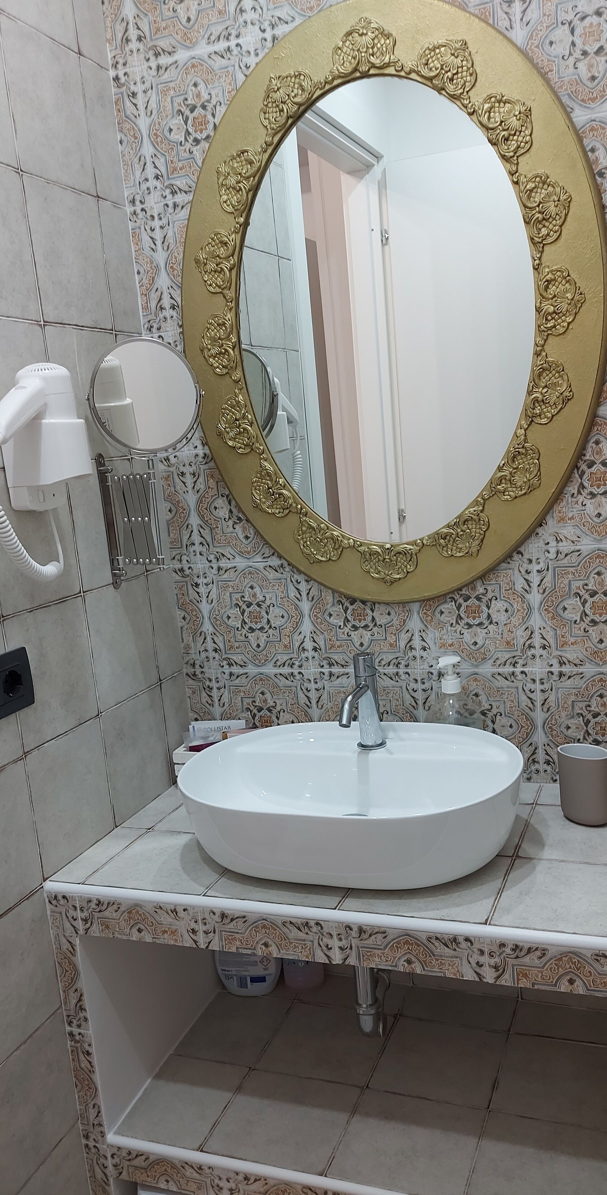 Un baño con un espejo redondo con marco dorado, un lavabo blanco y un secador de pelo.