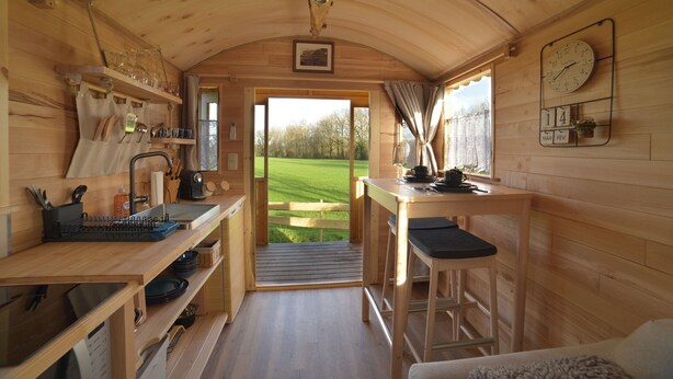 L’intérieur d’un chalet en bois comprend un coin cuisine, une table à manger avec un tabouret de bar, et une vue sur un champ verdoyant par une porte ouverte.