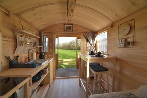 A wooden cabin interior with a kitchen area, a dining table with a bar stool, and a view of a green field through an open doorway. - Gîte insolite ''La Colibri''. A trailer in the heart of nature. (Chimay)