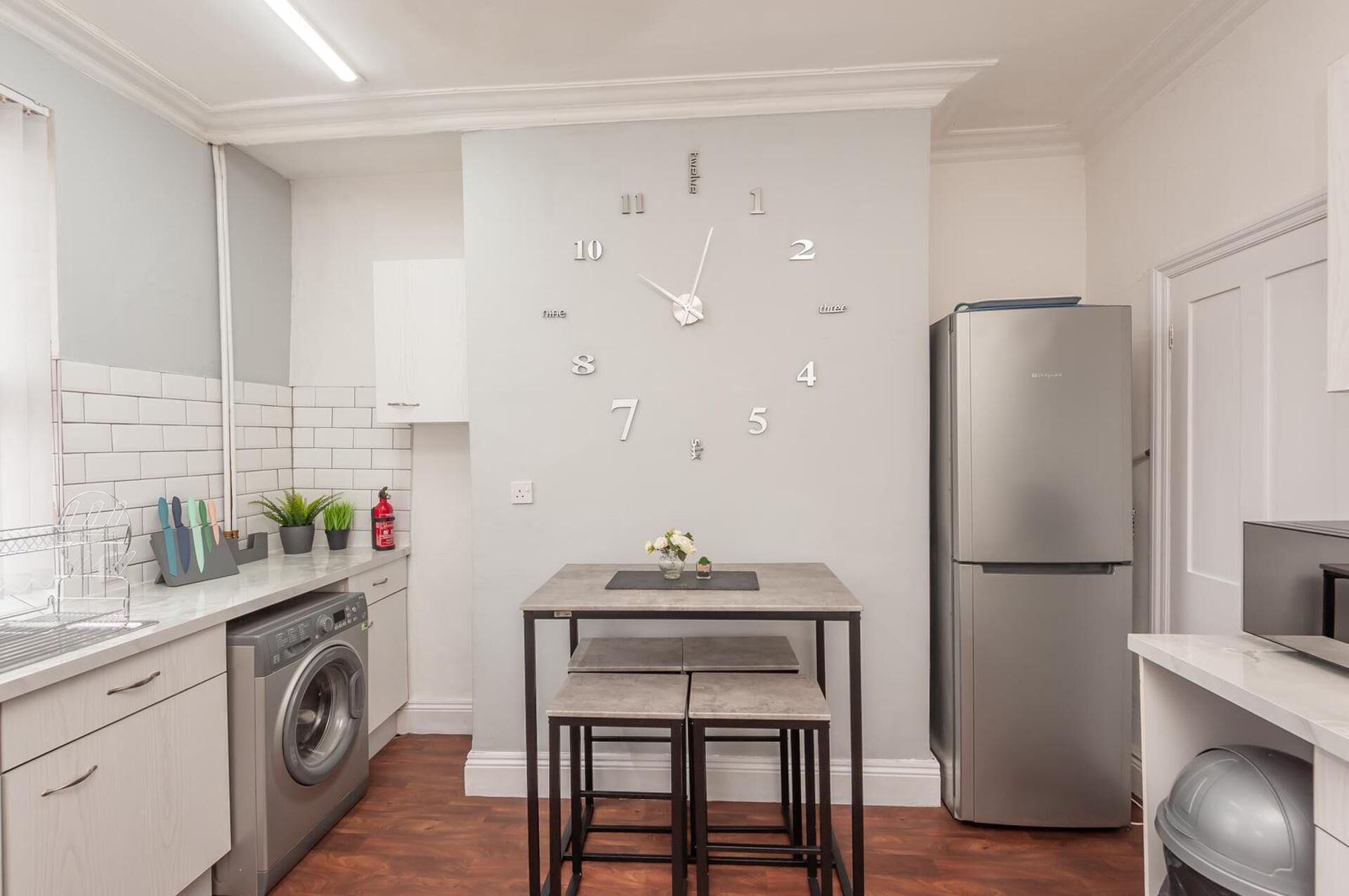 Eine Küche mit eingebauter Uhr, einer Waschmaschine, einem Kühlschrank und einem Esstisch mit Hocker.