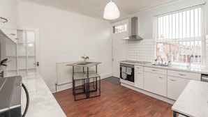 Una cocina moderna con armarios blancos, horno empotrado, campana extractora de acero inoxidable, ventana con persianas y una lámpara colgante.