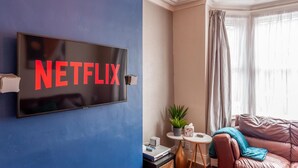 Sala de estar con un televisor montado en una pared azul que muestra 'NETFLIX', una mesita redonda con una planta, un sofá de cuero y una ventana con cortinas.