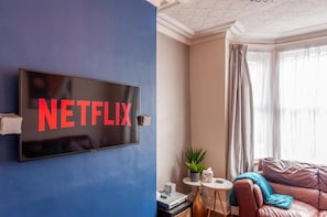 A living room with a TV mounted on a blue wall displaying 'NETFLIX,' a small round table with a plant, a leather sofa, and a window with curtains. - Leeds Lovely Home, 4 Beds Sleeps 9,free Parking (Leeds)