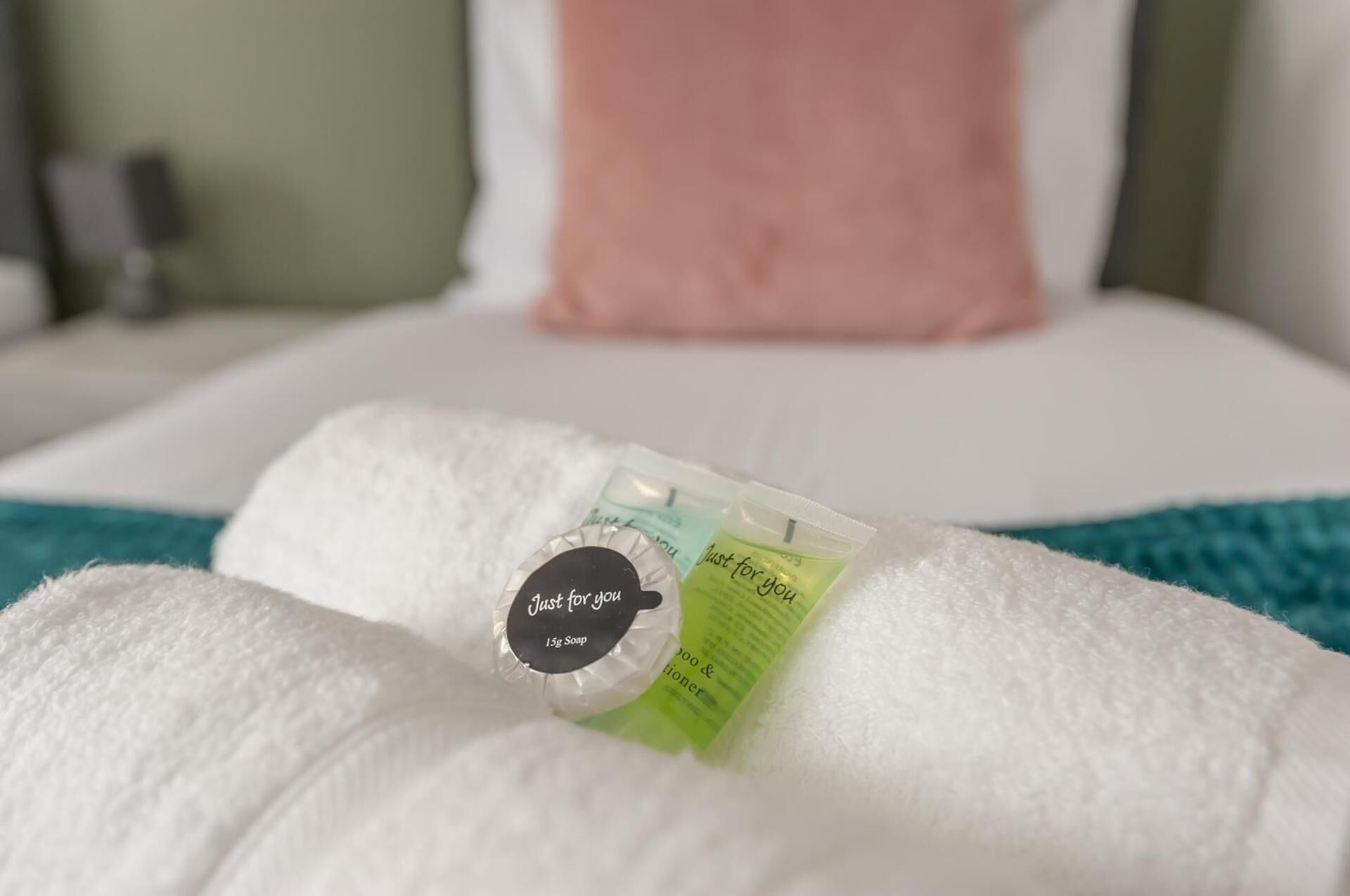 A neatly made bed with a white comforter, a pink pillow, and a small package of soap on the bed.