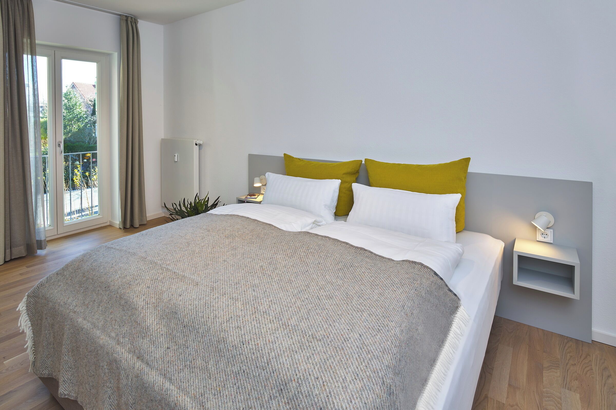 A bed with white bedding and two mustard pillows, a bedside table with a lamp, and a view of a balcony.