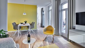 A modern living room with a dining area, a yellow accent wall, a large window with curtains, and a flat-screen TV on a white cabinet.