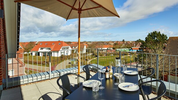 A balcony with a table set for dining, a large umbrella, and a view of houses in the background.