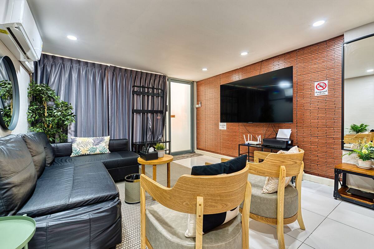 Un salon moderne avec un canapé en cuir noir, des chaises en bois, un téléviseur à écran plat et un mur décoratif en briques.