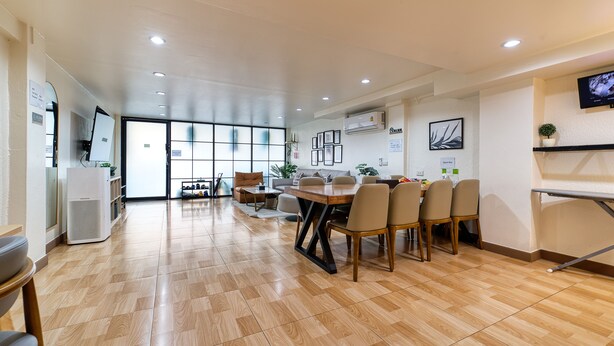 A spacious, well-lit room with a wooden floor, a dining area with a table and chairs, a TV mounted on the wall, and a large window providing natural light.