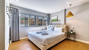 A modern bedroom with a large bed, a window with curtains, a small table with a plant, and a wall-mounted lamp.