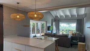 Een moderne keuken met een wit eiland en gouden handgrepen, een eethoek met tafel en stoelen, een zithoek met een bank en televisie, en een balkon met uitzicht op groen.