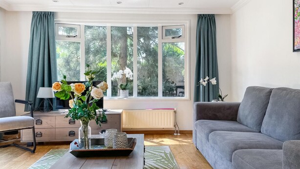 A living room with a grey sofa, a wooden coffee table with decorative items, a desk with a chair, and a large window with curtains. - Stunning (sta)caravan in Putten (Putten)