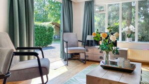A modern living room with a sofa, armchair, and a coffee table. There is a TV on a stand, a vase with flowers, and a window with curtains. - Stunning (sta)caravan in Putten (Putten)