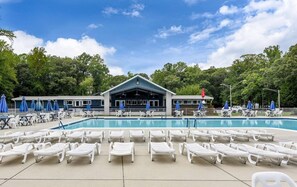 Une piscine entourée d’arbres et d’un bâtiment doté d’une zone abritée, avec des chaises longues blanches et des parasols bleus.