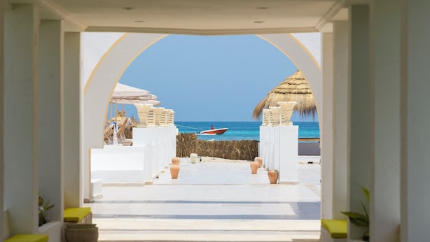 Une vue sur la plage, encadrée par une porte en arc, avec un bateau sur l’eau, des plantes en pot et une structure au toit de chaume.