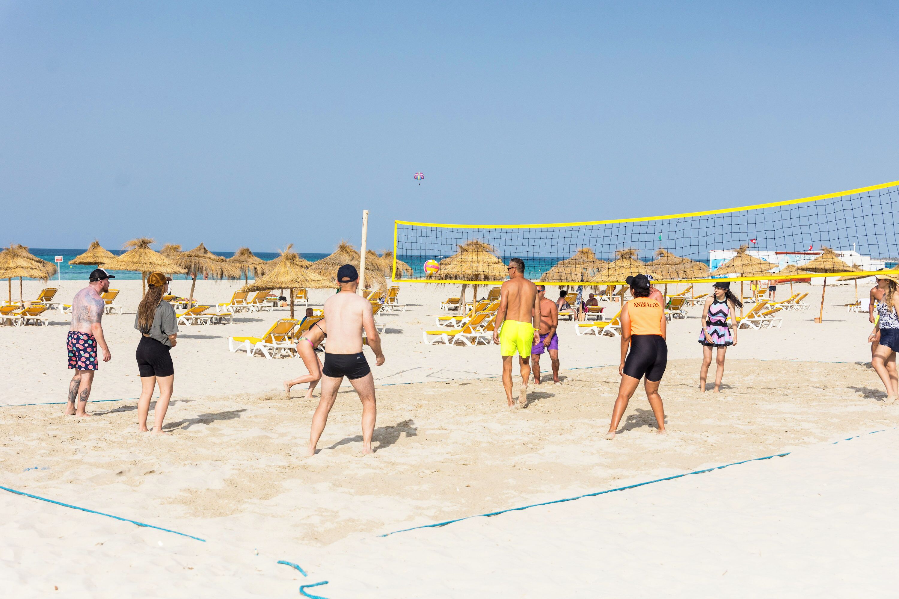 A beach volleyball game with players and spectators, sun loungers, and thatched umbrellas.