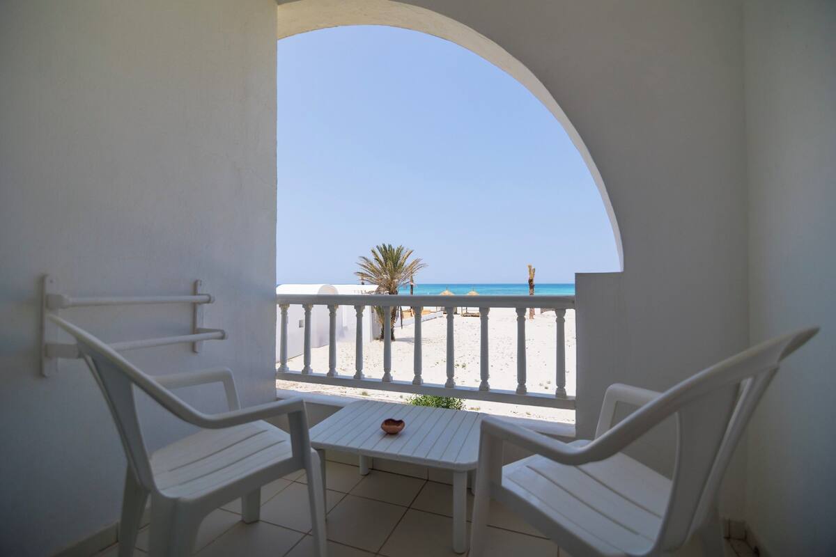En balkon med hvide stole og et lille bord, med udsigt over en strand med palmetræ og parasoller.