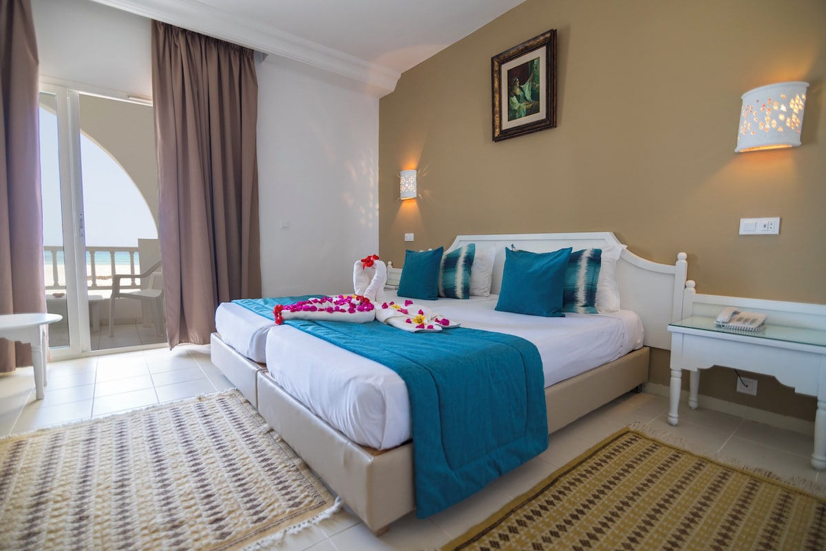A hotel room with a bed, a bedside table, a lamp, a picture on the wall, and a view of the outside.
