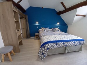 Ein Schlafzimmer mit schrägen Wänden, einem Bett mit gemusterten Textilien, einem Holzschrank und einem Hocker.