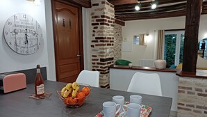 A dining area with a wooden table, a fruit bowl, a bottle, and white chairs. - Les Maisons du Théâtre Romain - 4 Gîtes - 10 Chambres (Le Vieil-Évreux)