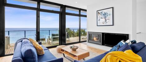 A modern living room with a large window overlooking the sea, a fireplace, and a cozy seating area.