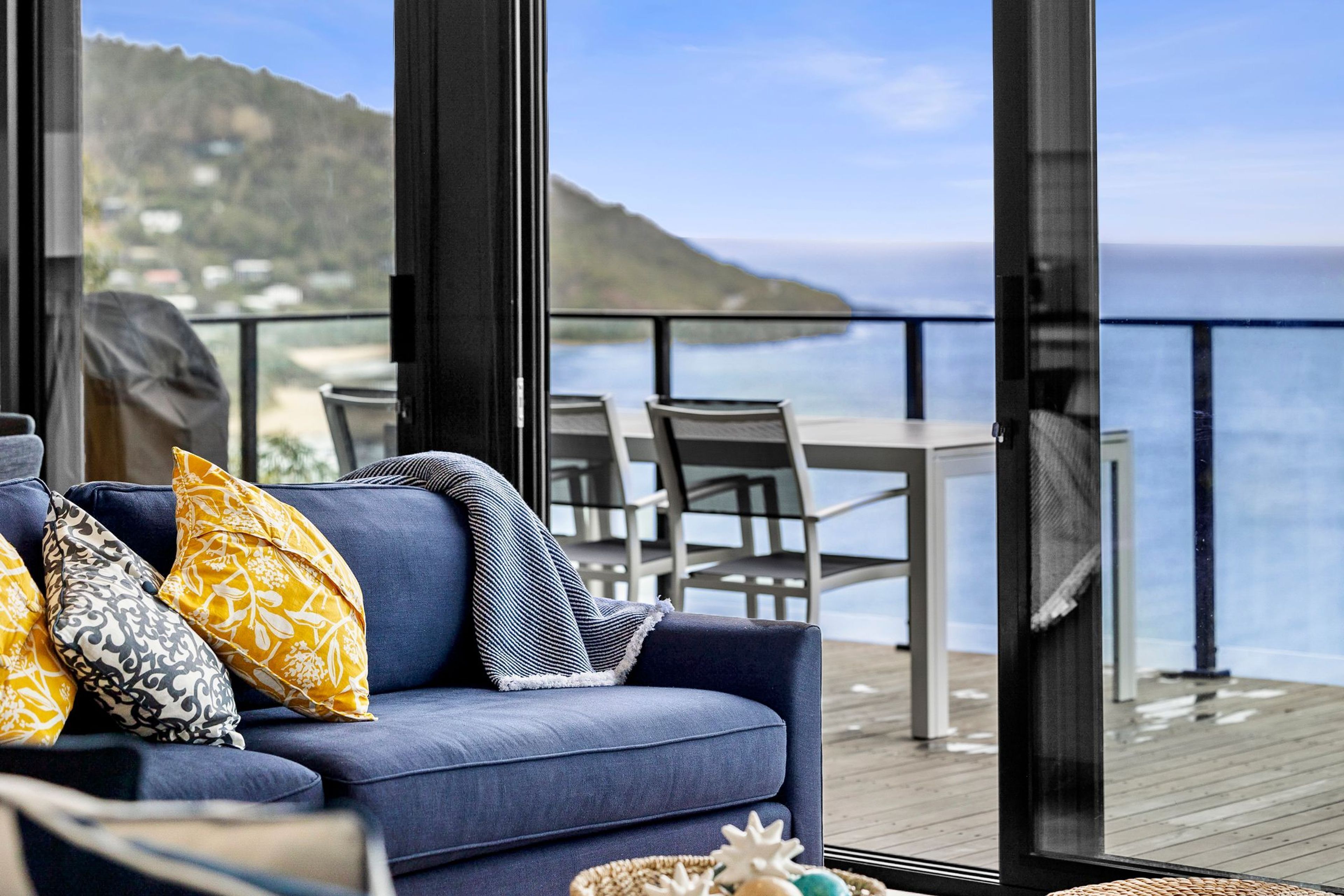 Ein modernes Wohnzimmer mit einem blauen Sofa, gemusterten Kissen und Meerblick durch große Fenster.