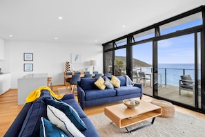 Ein modernes Wohnzimmer mit einem großen Fenster mit Meerblick, einem Essbereich mit einem Holztisch und einer gemütlichen Sitzecke mit einem blauen Sofa und Kissen.