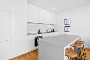 A modern kitchen with white cabinetry, a grey countertop, and a built-in oven. There are two bar stools with wooden seats and metal legs. - Wonder Wye (Wye River)