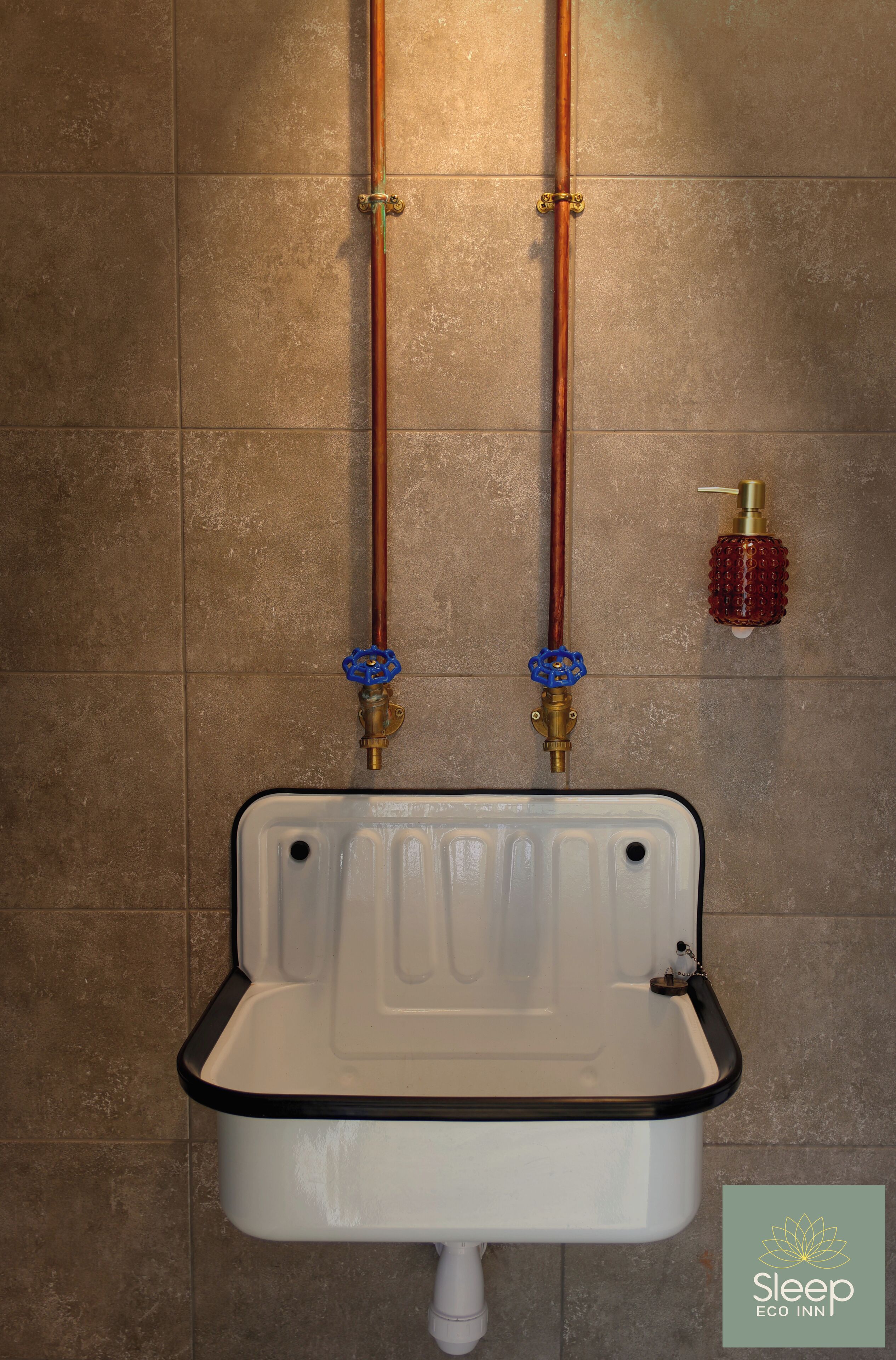 Un baño con un lavamanos blanco, dos tuberías de cobre y un dispensador de jabón rojo.