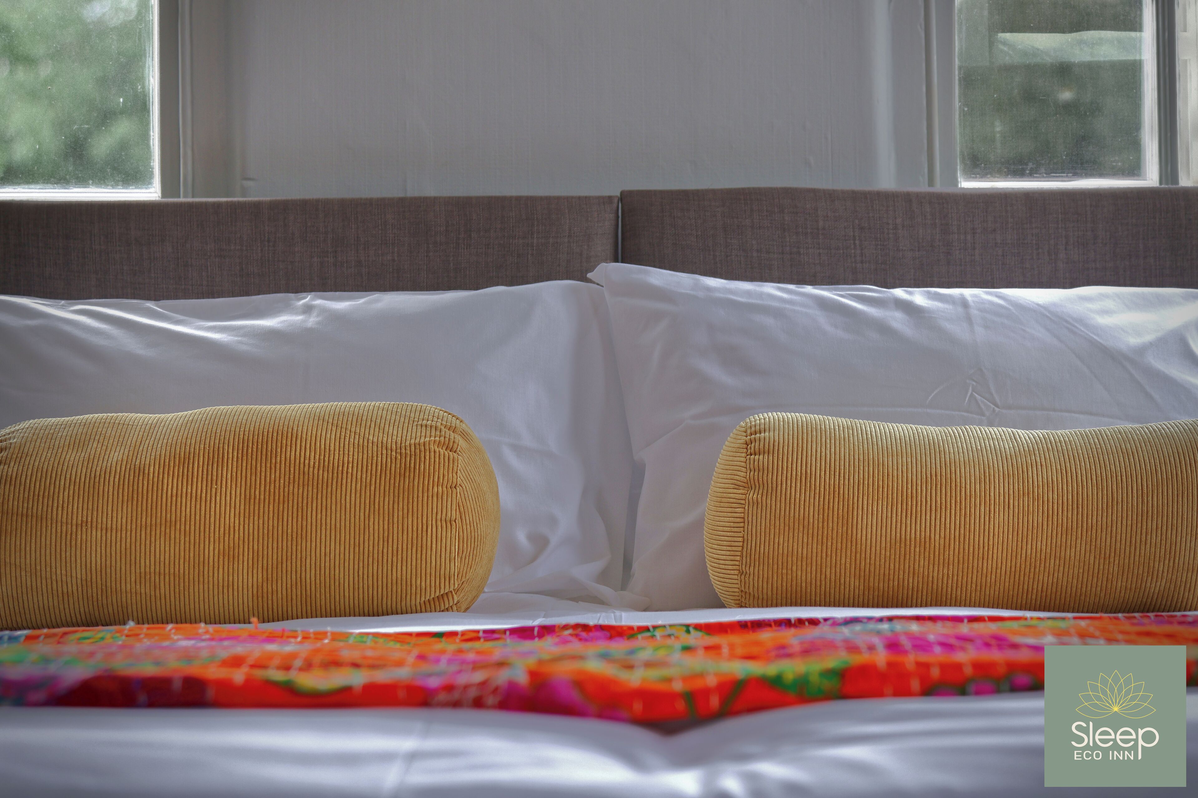 Un lit avec du linge de lit blanc et deux oreillers beige, une courtepointe aux couleurs vives et une fenêtre donnant sur des arbres.