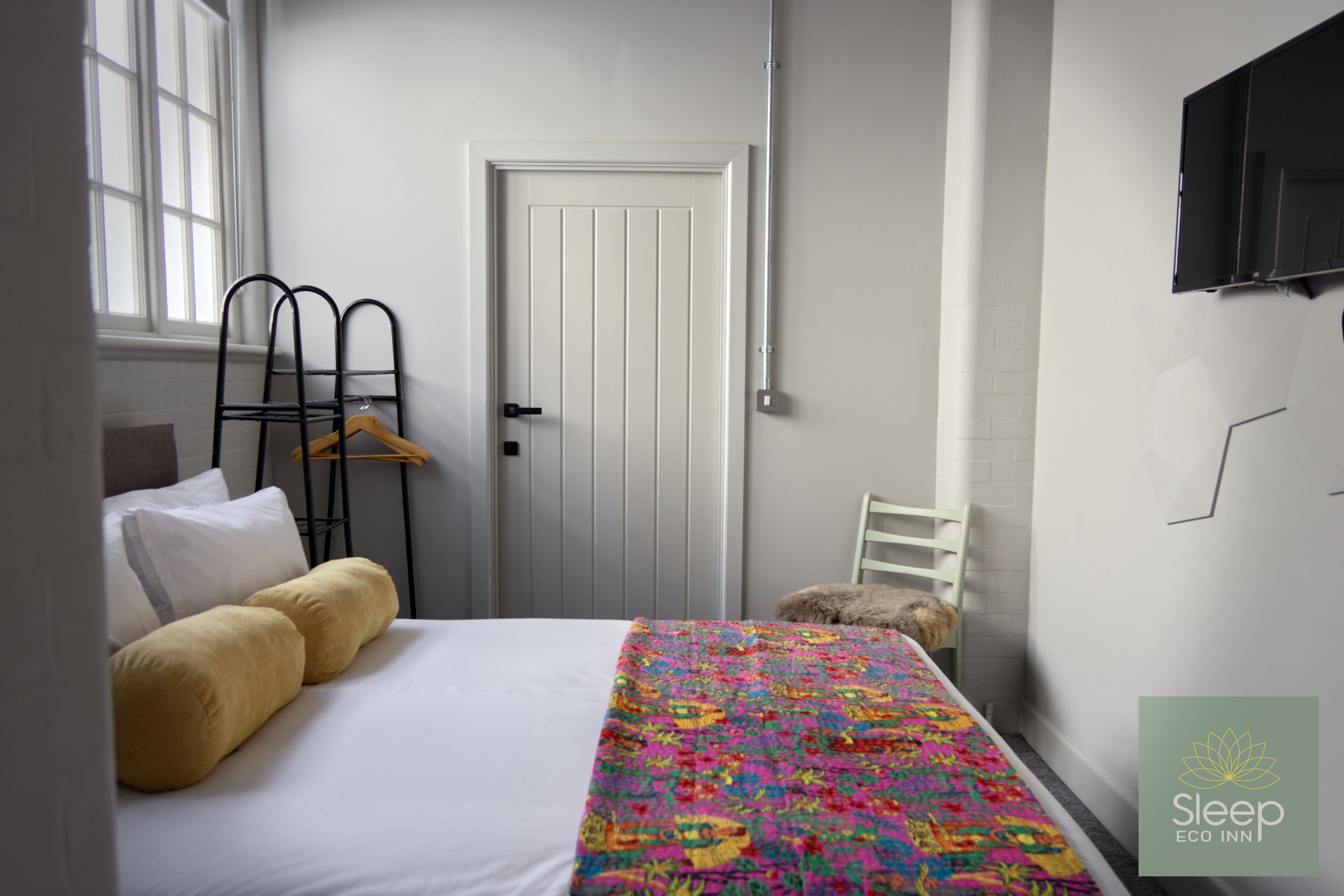 A bedroom with a bed, a colorful quilt, a white door, a coat rack, and a TV on the wall.