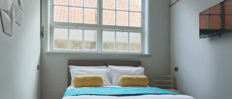 A bed with white bedding and two yellow pillows, a window with a view of a brick building, and a wall-mounted TV.