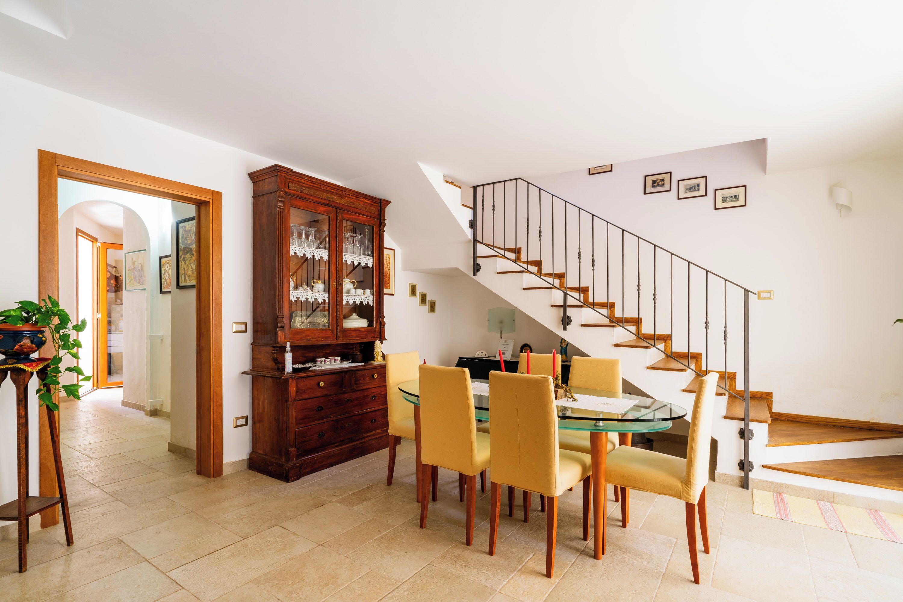 A dining area with a glass table and yellow chairs, a wooden cabinet with glass doors, and a staircase with a metal railing.