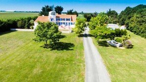 Une grande maison avec une allée pavée, entourée de champs verdoyants et d’arbres.