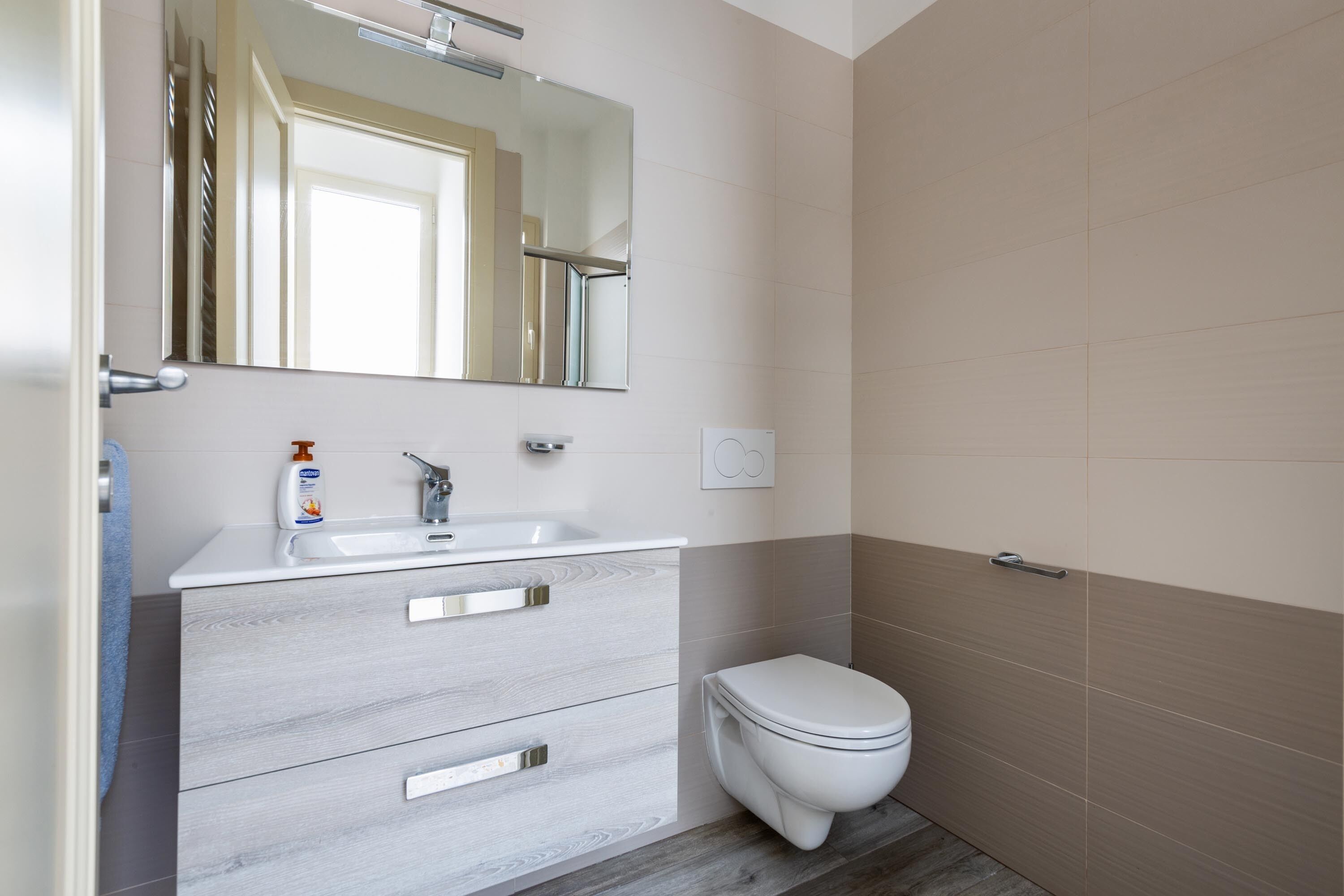 Une salle de bain moderne avec un meuble-lavabo blanc, un miroir, des toilettes et des murs recouverts de carreaux beiges.