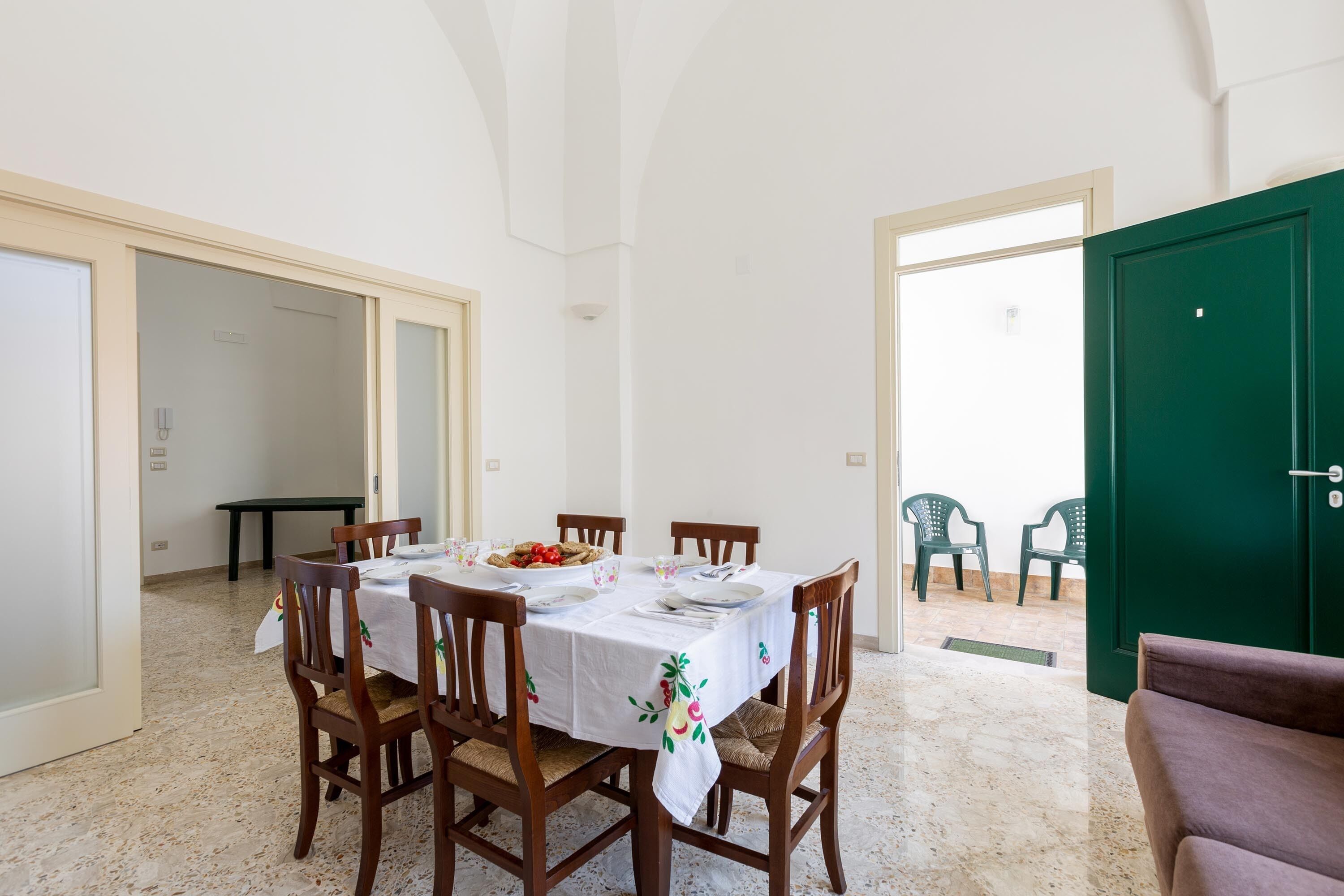Un comedor con una mesa dispuesta para cuatro personas, rodeada de sillas de madera. Se ve una puerta verde y un banco al fondo.