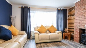 A living room with a beige sofa, a smaller sofa with yellow cushions, a brick fireplace, and a wooden floor. - 17 Third Avenue (Prestatyn)