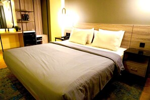 A hotel room with a large bed, a bedside table with a lamp, a small desk with a computer, and a shelf with a coffee maker. - Series Hotel Sudirman (Jakarta)