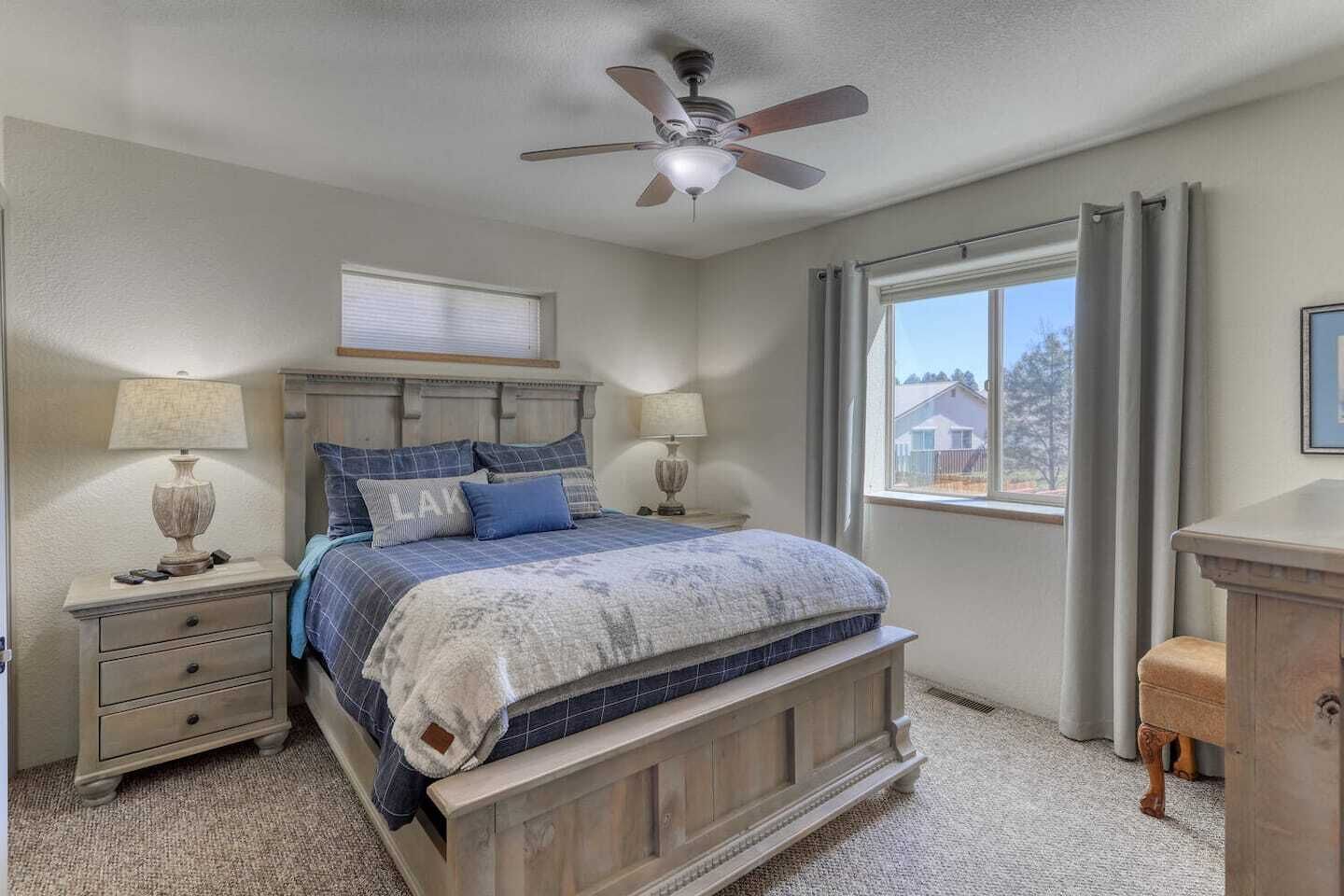 A bedroom with a bed, two bedside tables, a ceiling fan, a window with curtains, and a view of houses outside.